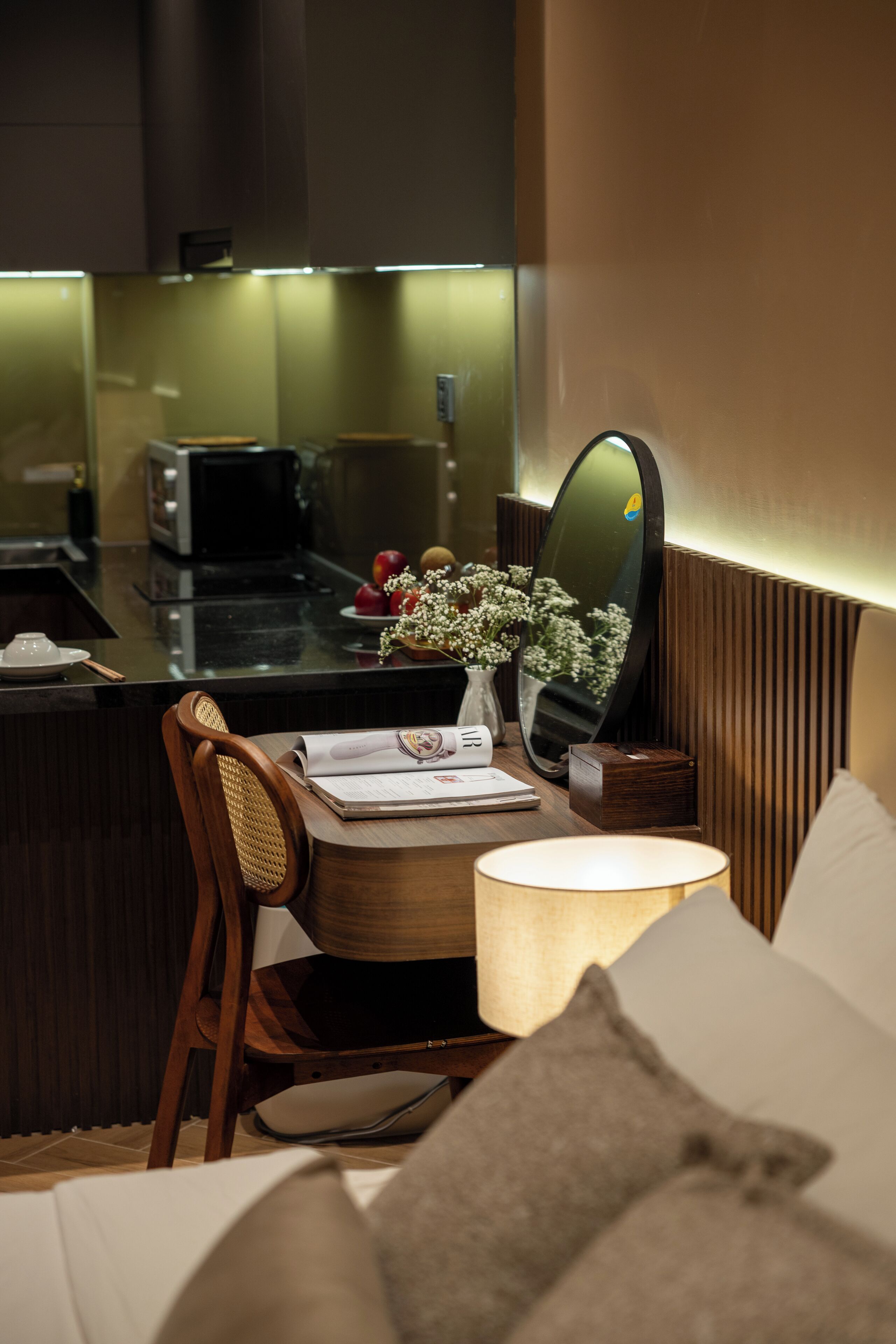 A modern hotel room with a wooden chair, a bedside table with a lamp, a mirror, and a view of the kitchen area.