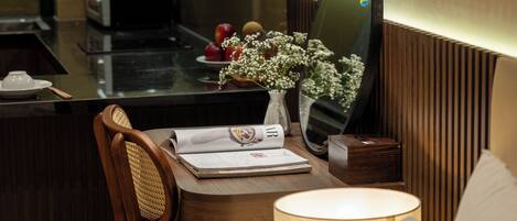 A modern hotel room with a wooden chair, a bedside table with a lamp, a mirror, and a view of the kitchen area.