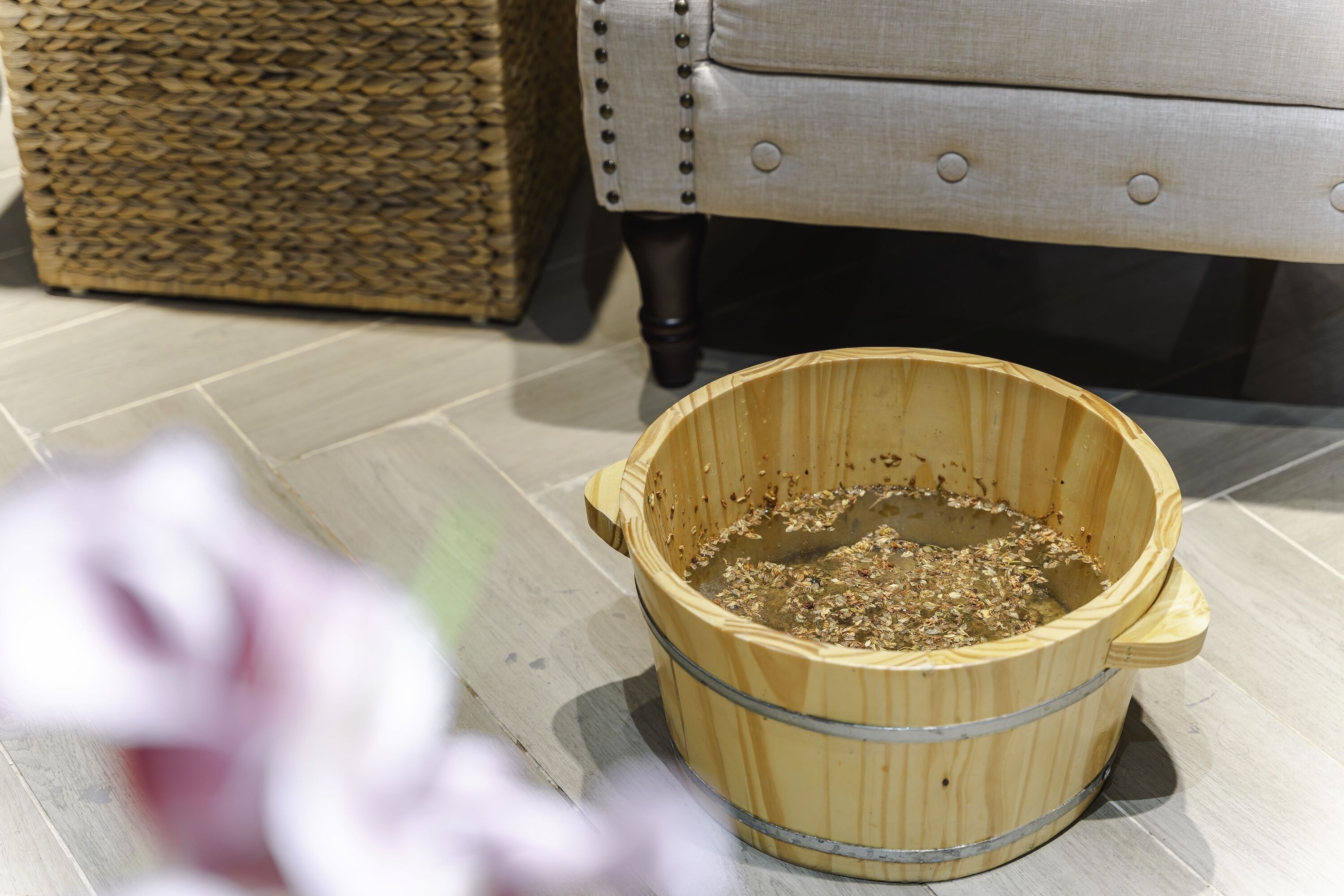 A wooden bucket on a tiled floor with a wicker basket and a cushioned chair in the background.