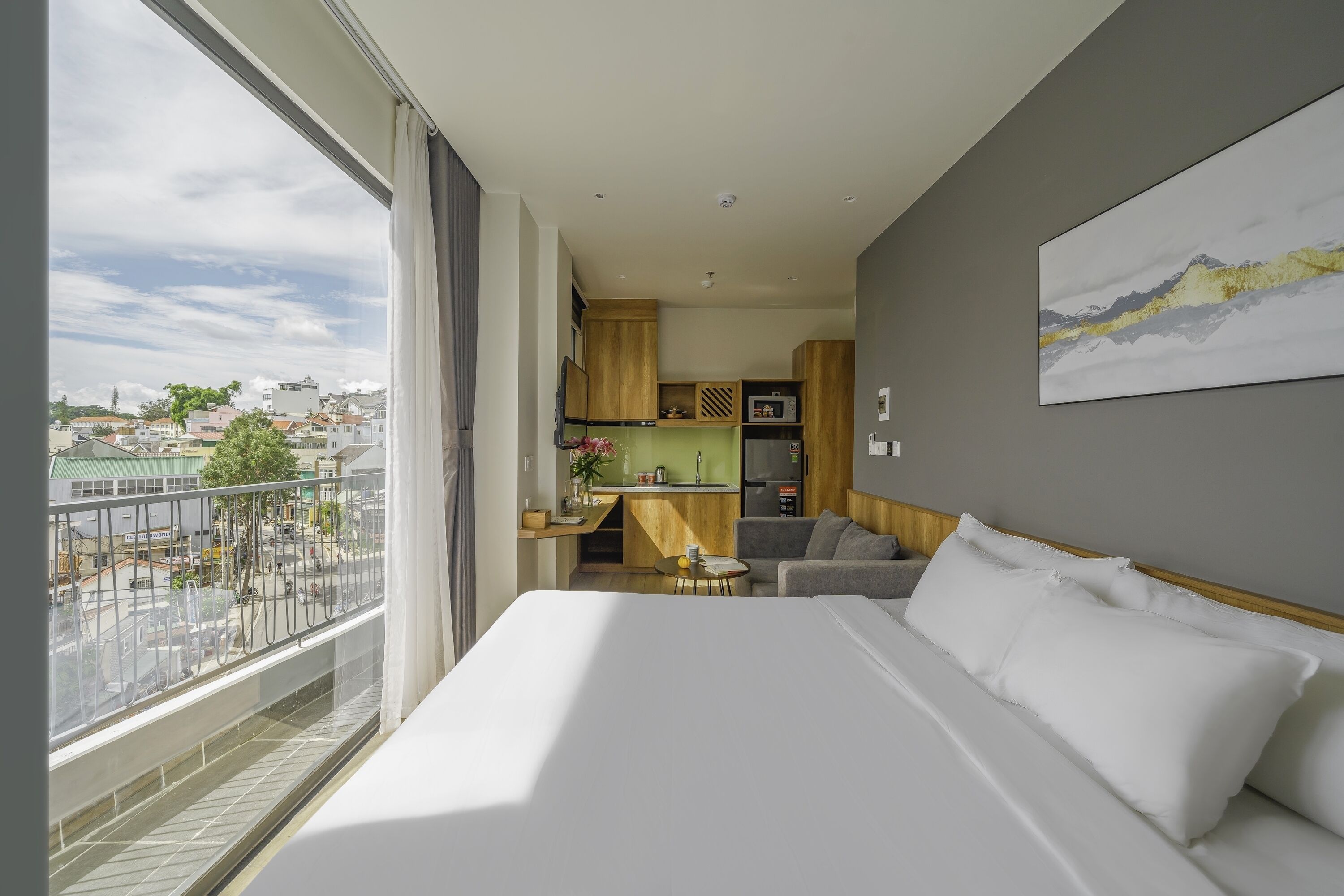 A hotel room with a large bed, a small sofa, a kitchen area, and a balcony with a city view.