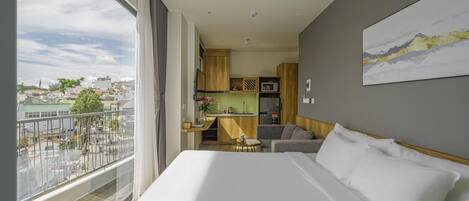 A hotel room with a large bed, a small sofa, a kitchen area, and a balcony with a city view.