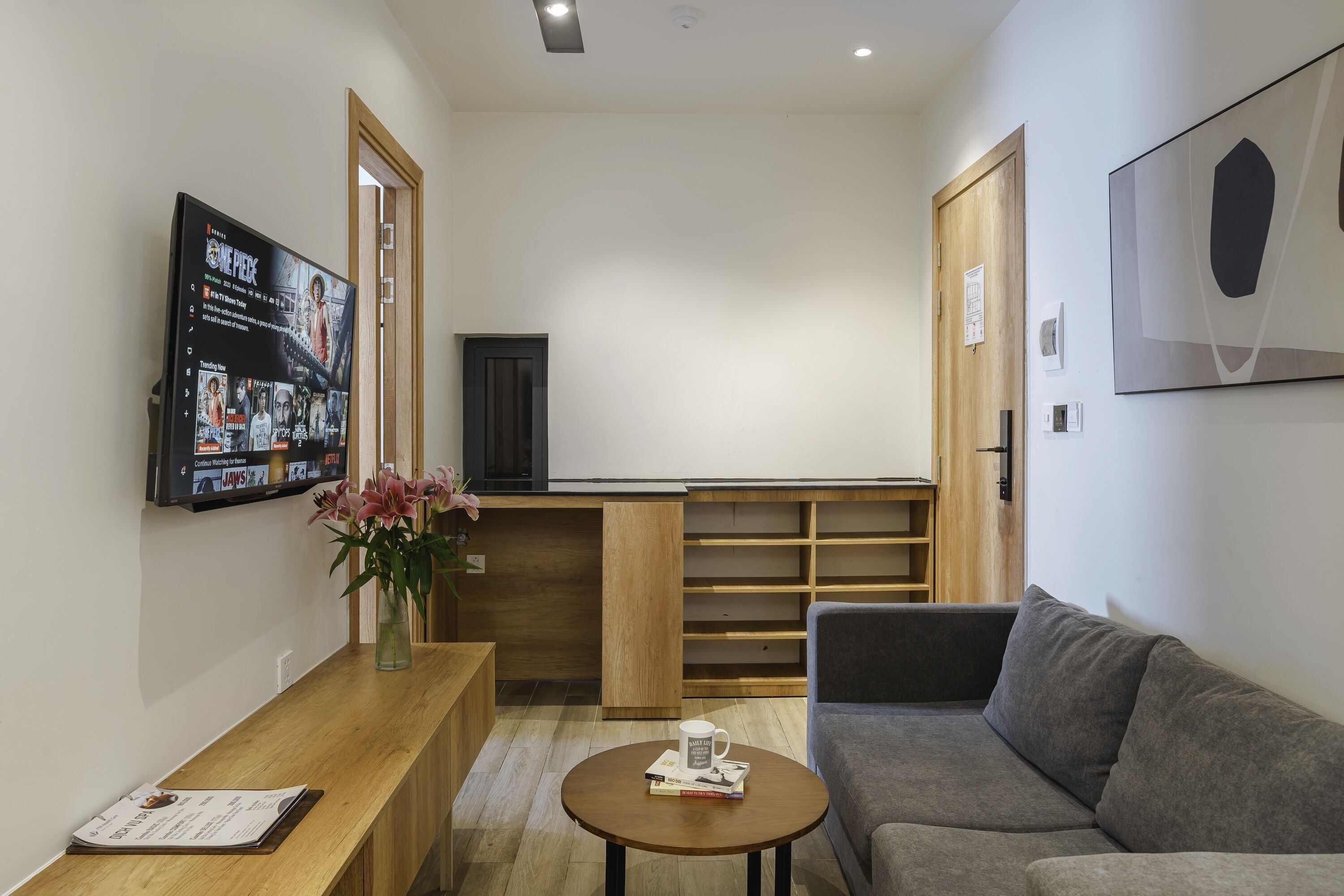 A modern living room with a grey sofa, a wooden coffee table, a TV mounted on the wall displaying a movie, and a wooden reception desk.