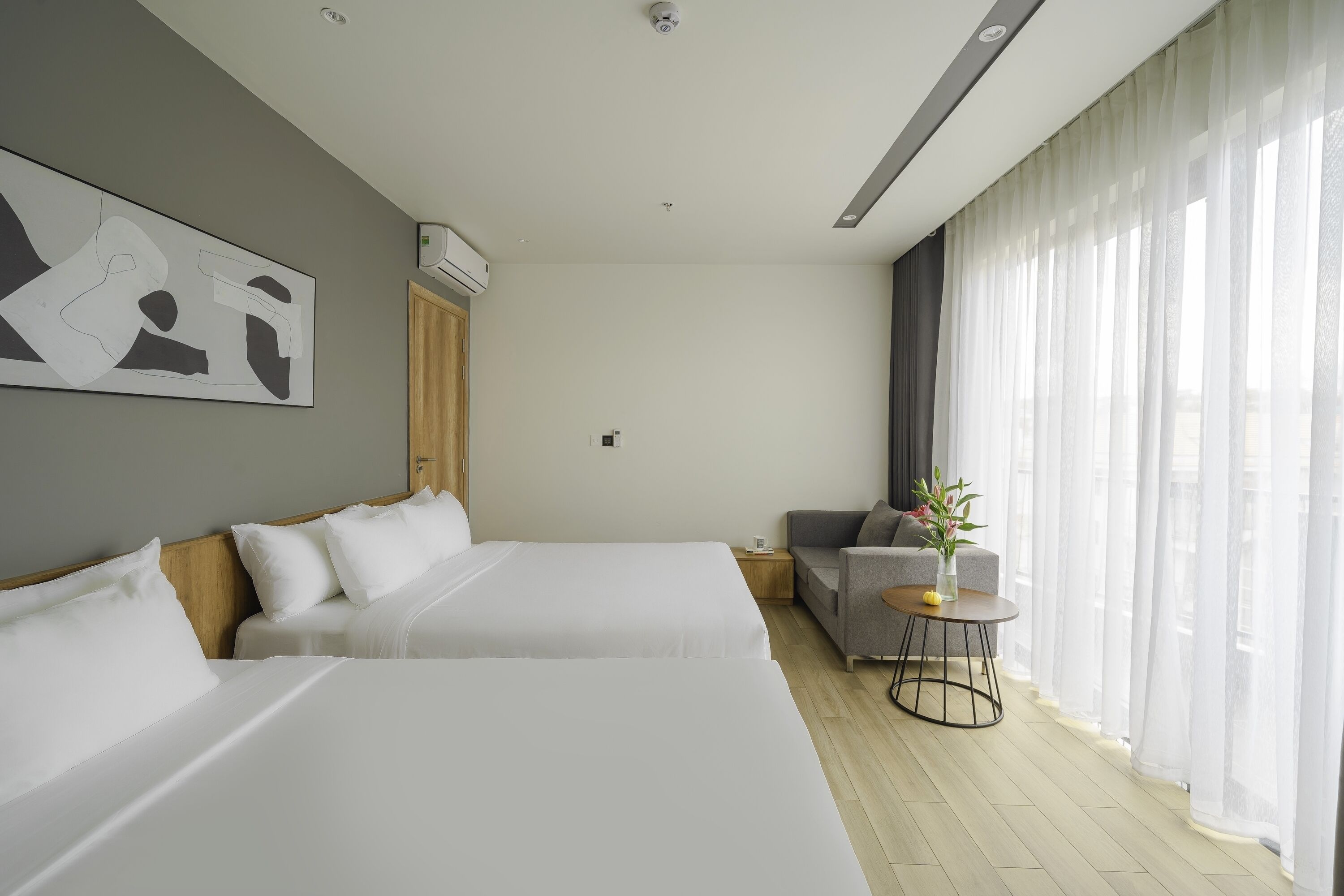 A modern hotel room with two beds, a sofa, a small table, and a large window with sheer curtains.