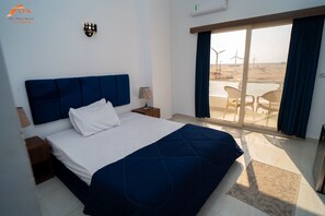 Une chambre à coucher avec un lit, une table de chevet, une lampe et une vue sur la plage, où l’on aperçoit des éoliennes.