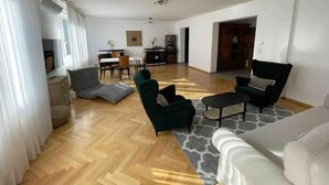 Un salon spacieux doté d’un parquet en bois disposé en chevrons, d’un canapé, de deux fauteuils et d’une table basse. Au fond, on aperçoit un coin repas avec une table et des chaises.