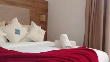 Habitación de hotel con cabecera de madera, ropa de cama blanca, manta roja y un juego de toallas blancas.