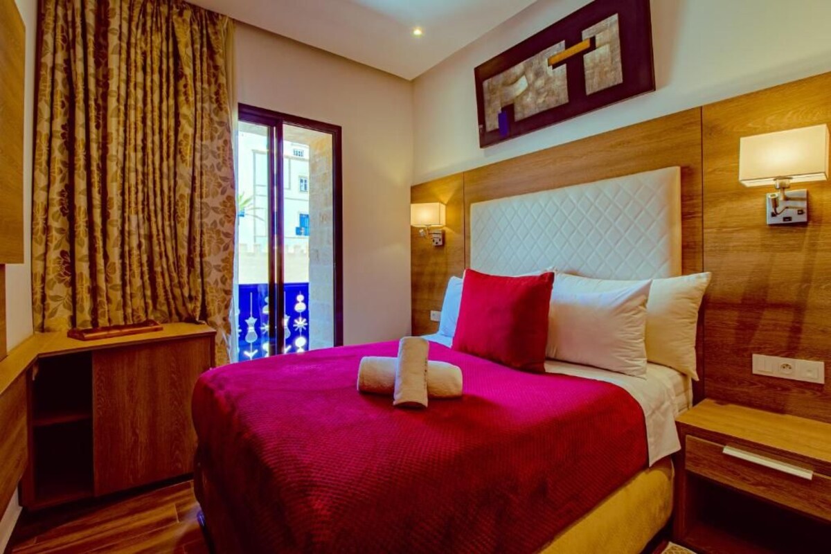 A hotel room with a large bed, wooden headboard, a nightstand, a lamp, and a view of the outside.