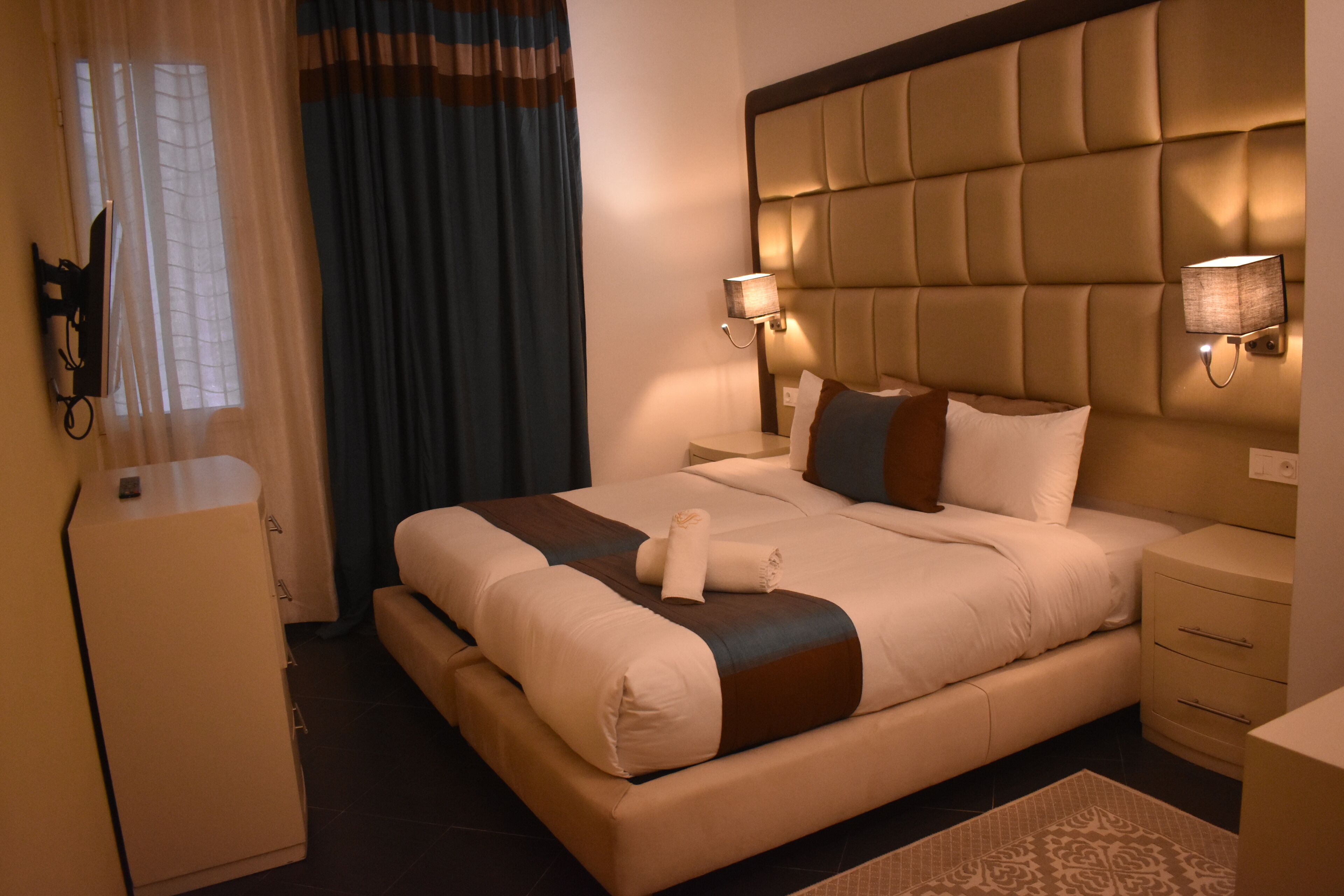 Una habitación de hotel con una cama grande, dos mesitas de noche, un armario y una ventana con cortinas.