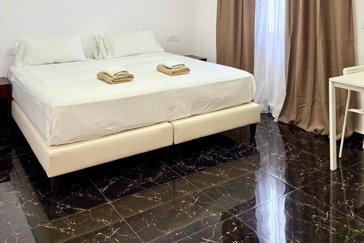 Een hotelkamer met een groot bed, een kroonluchter, een bureau en uitzicht op het landschap door gordijnen.
