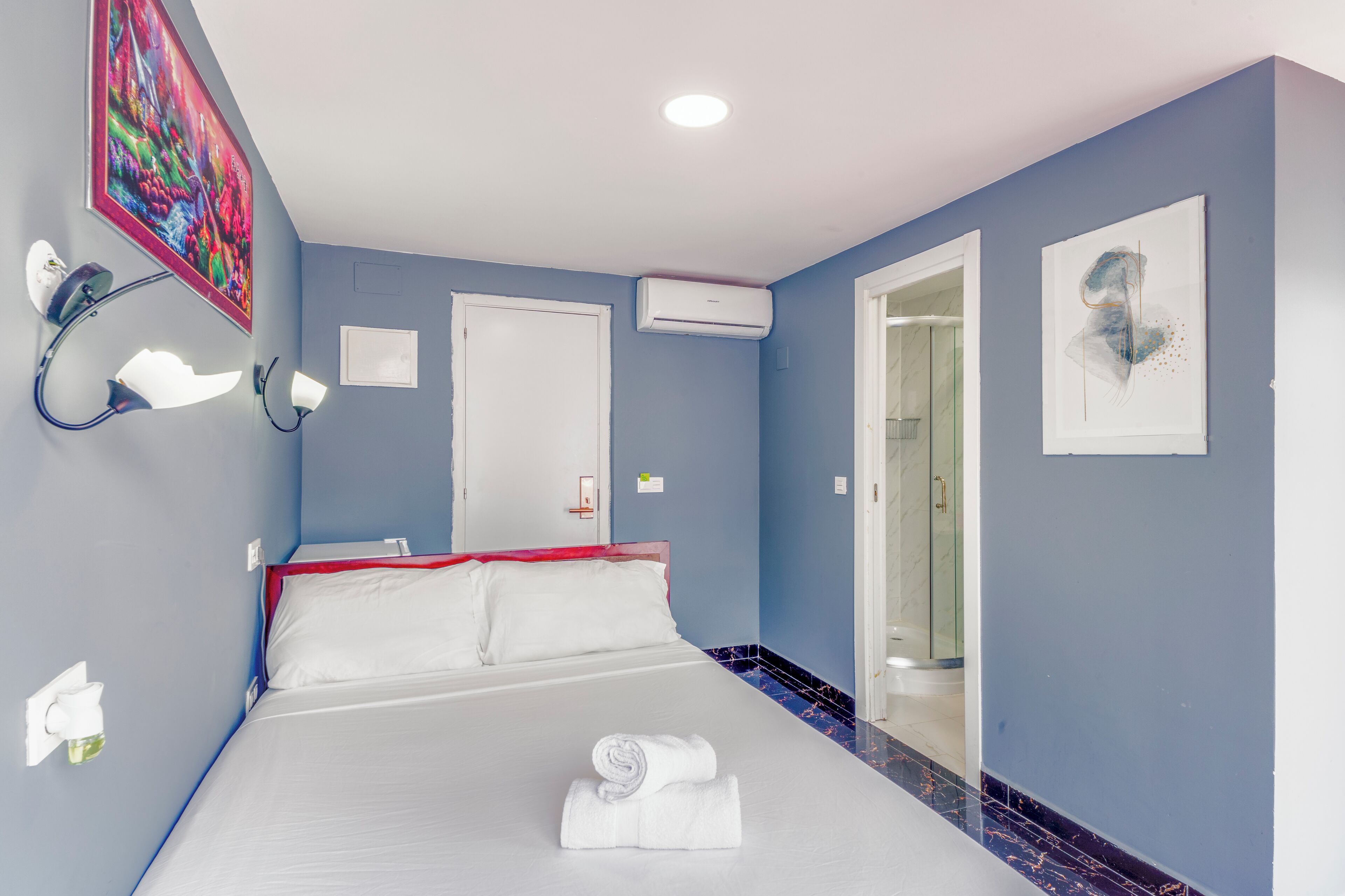 A hotel room with a bed, a door, an air conditioning unit, and a bathroom visible through an open door.