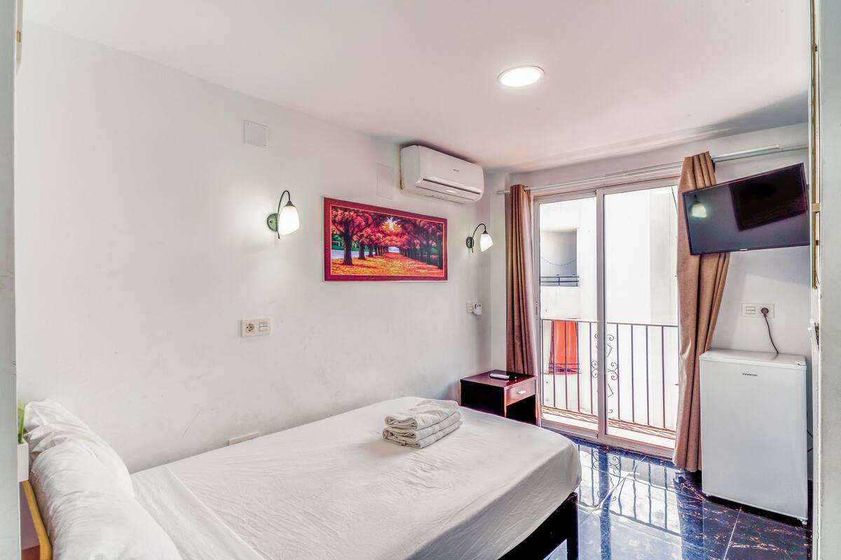 Een hotelkamer met een bed, een bureau, een airconditioner, een televisie en een balkon met gordijnen.