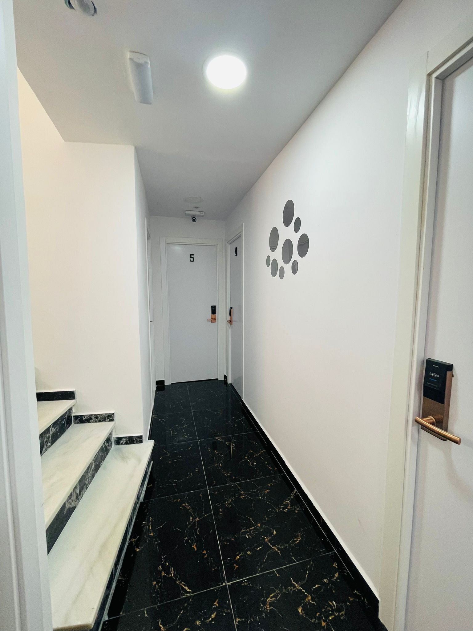 Un pasillo con una escalera de mármol, una puerta blanca con un número y una pared decorada con motivos circulares.