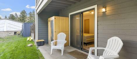 A covered patio with two Adirondack chairs, a sliding glass door leading to a bedroom, and a small outdoor area with a shed and bags.