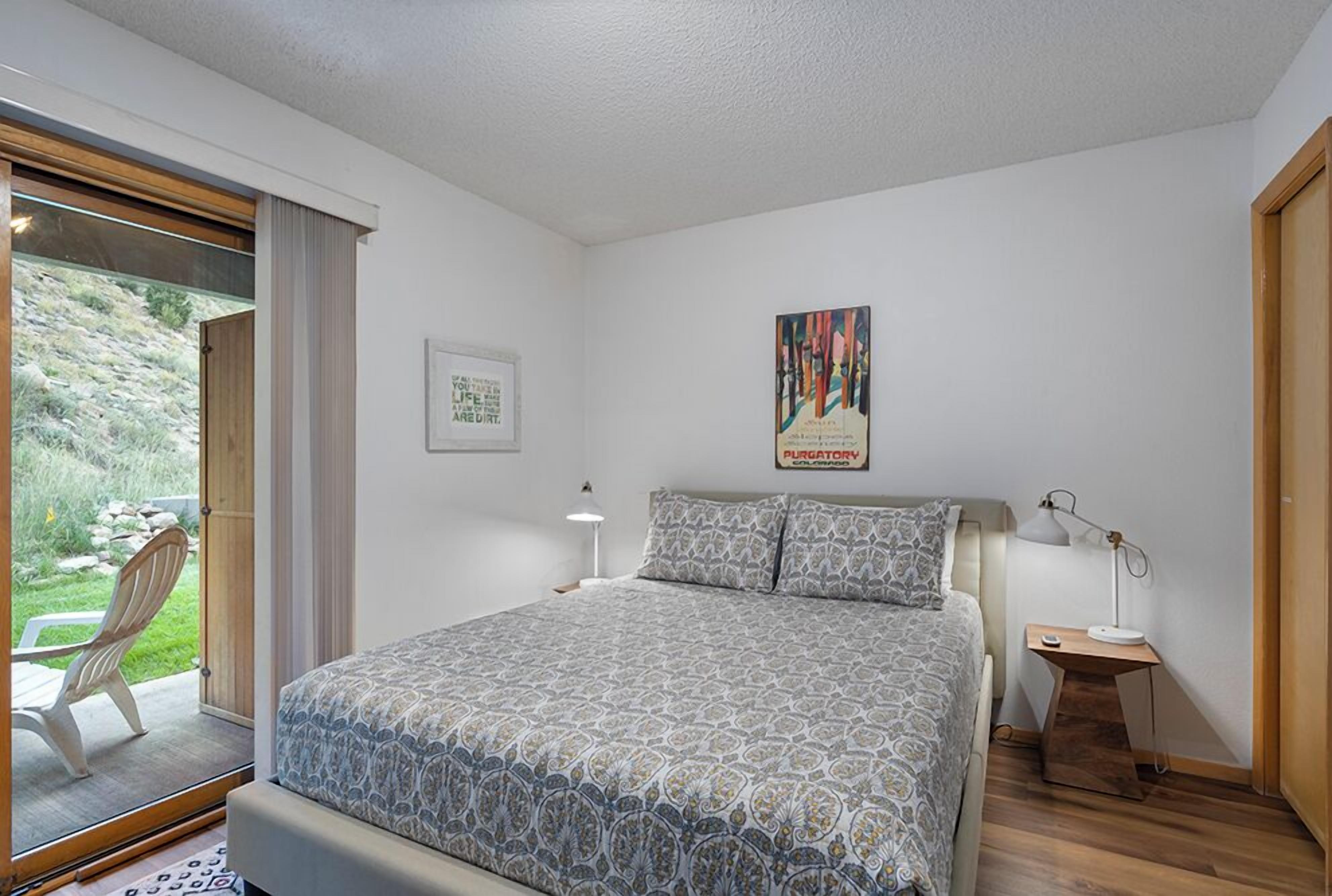 Une chambre à coucher avec un lit, une table de chevet avec une lampe, un tableau encadré au mur et une fenêtre donnant sur un paysage verdoyant.