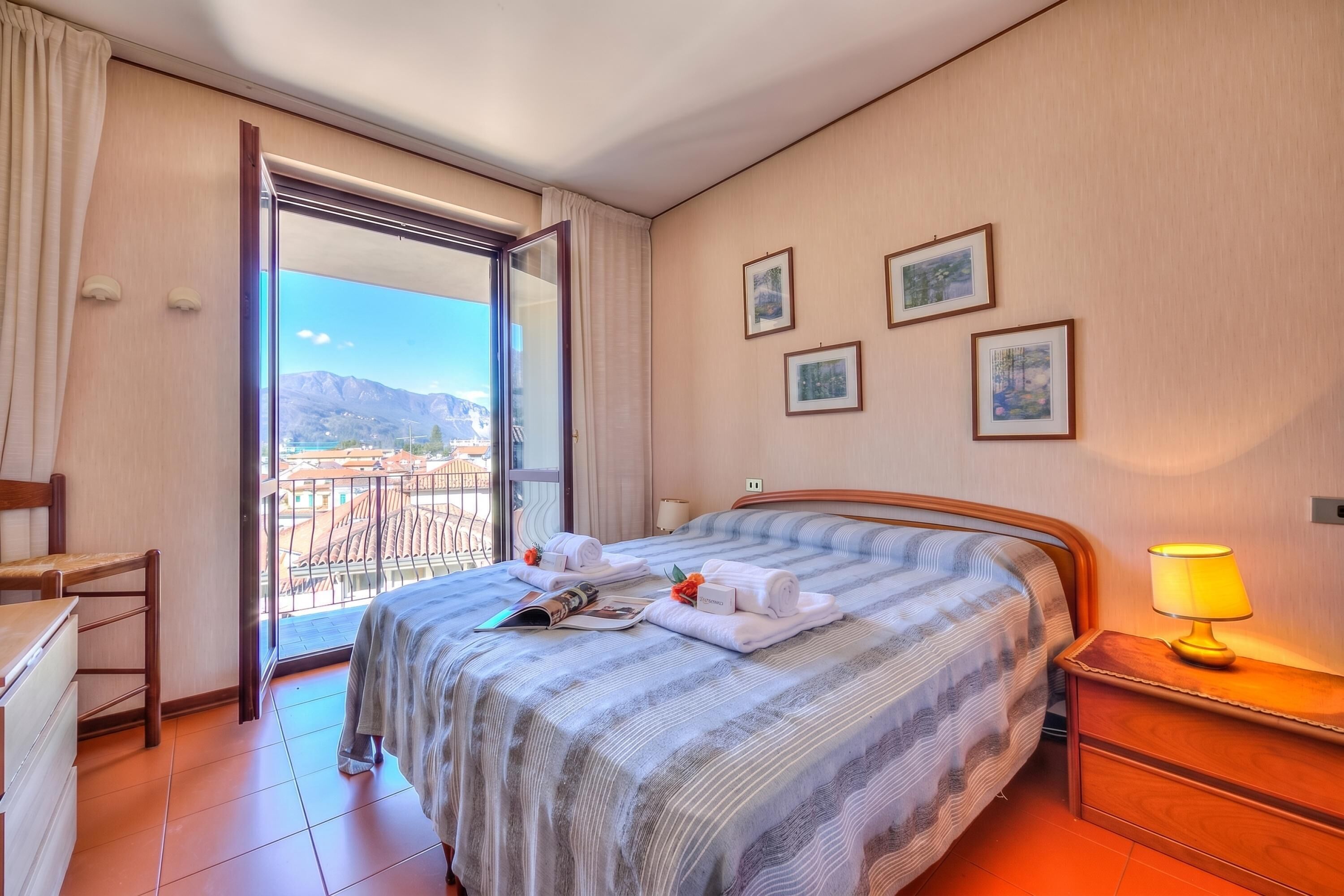 A hotel room with a bed, a bedside table with a lamp, a chair, a desk, and a balcony with a view of buildings and mountains.