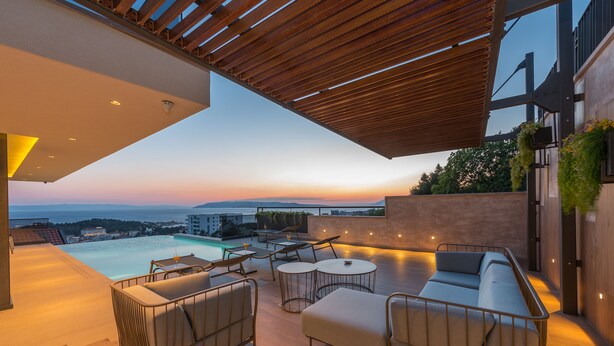 A modern outdoor lounge area with a pool, seating, and a view of the sunset over the ocean.
