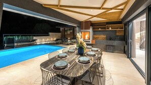 A modern outdoor dining area with a pool, a fire feature, and a kitchen area with a stone backsplash.