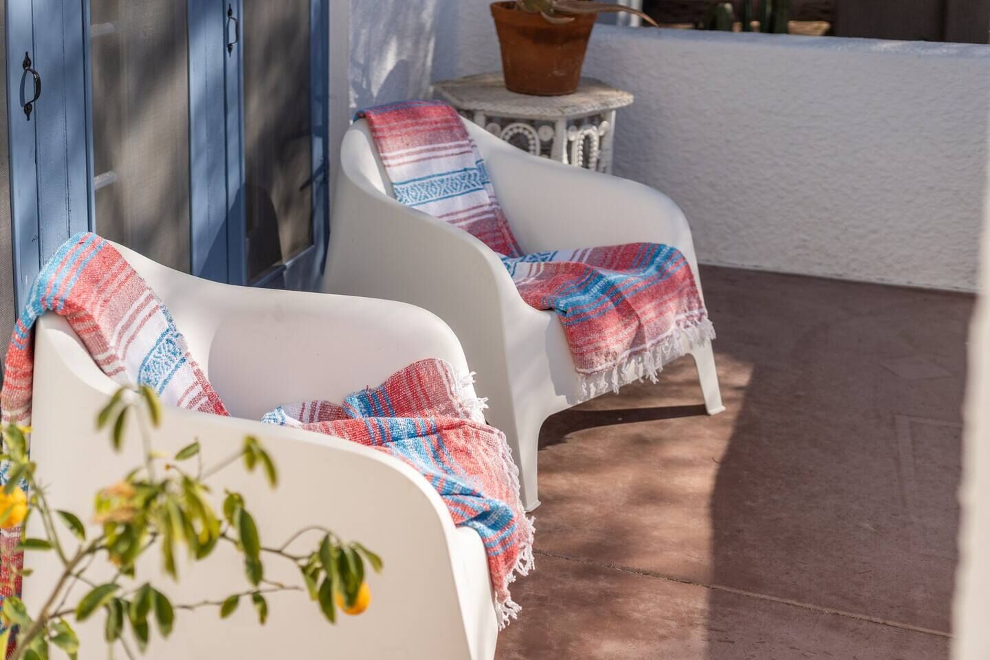 Two white patio chairs with colorful blankets, a potted plant on a small table, and a blue door in the background.