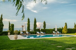 A pool area with lounge chairs and umbrellas, surrounded by greenery and tall trees.