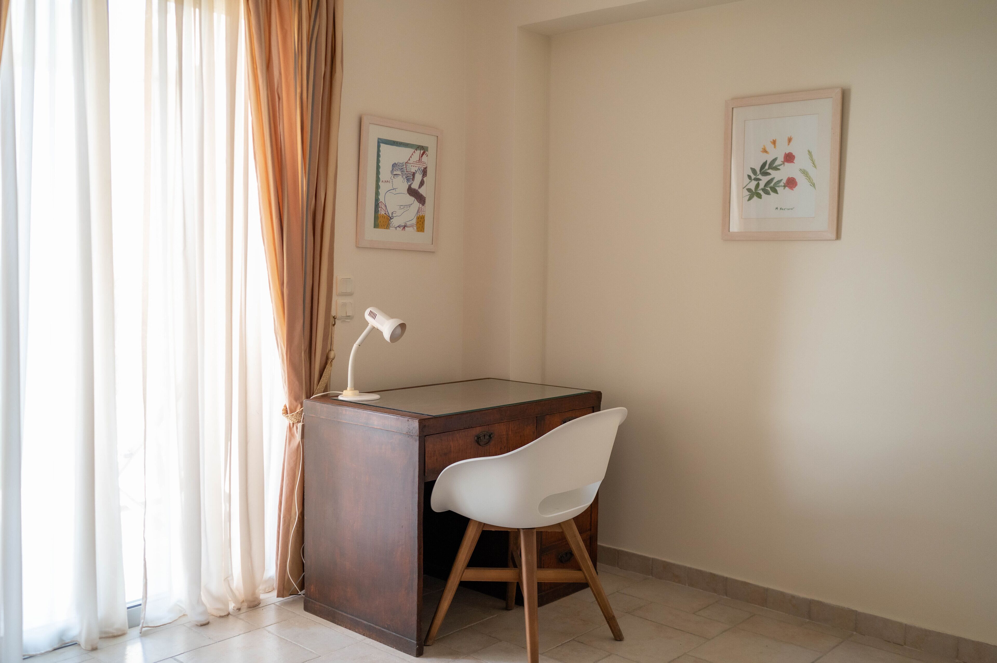 Une petite pièce bien éclairée, avec un bureau, une chaise et deux reproductions botaniques encadrées accrochées au mur.