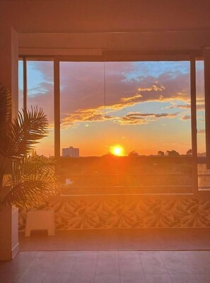Ein Zimmer mit großem Fenster und Blick auf einen Sonnenuntergang, einem kleinen Hocker und einer Topfpflanze.