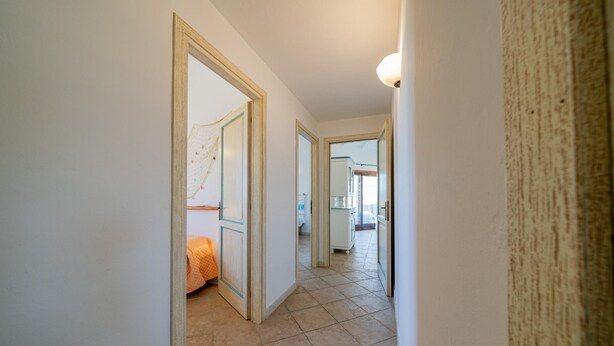 Un couloir étroit au sol carrelé, aux murs blancs, et une porte donnant sur une pièce où se trouve un lit orange.