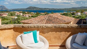 Un balcon agrémenté de fauteuils et de coussins en osier, offrant une vue sur une ville côtière aux toits de tuiles rouges et sur une mer d’un bleu limpide.