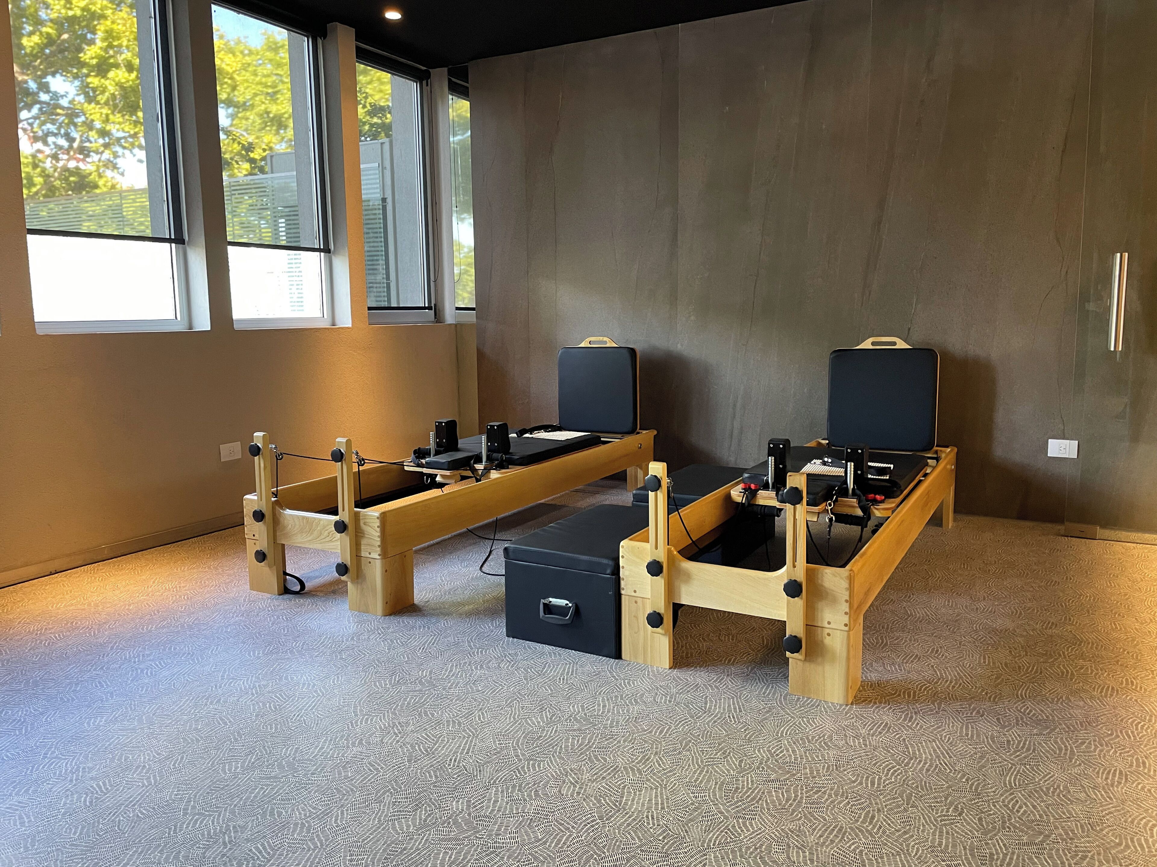 A room with two wooden massage tables, each with a black cushion and a small black speaker. The room has large windows, a carpeted floor, and a plain wall.
