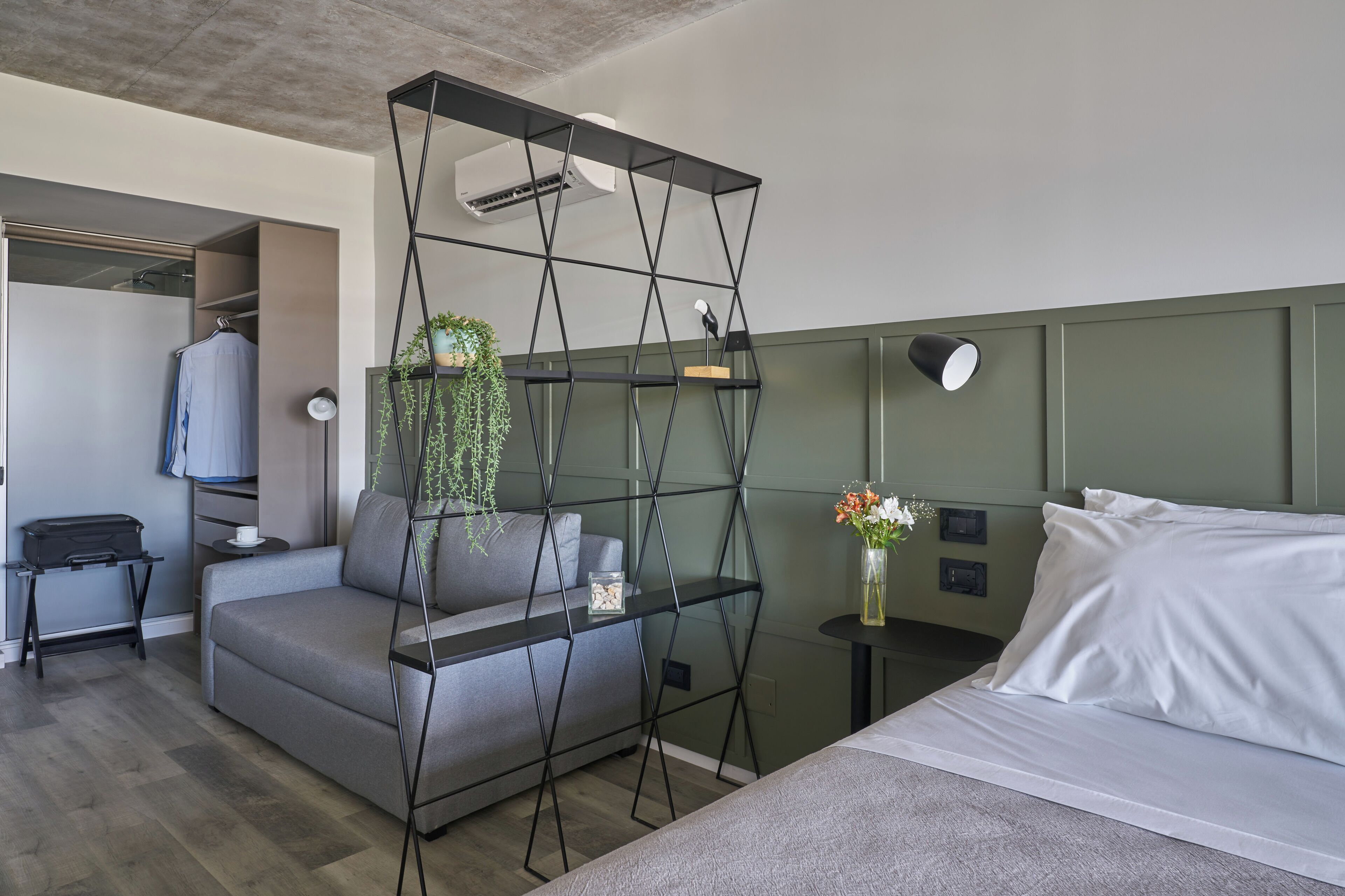 Ein modernes Hotelzimmer mit einer grauen Couch, einem Bett mit weißen Leinen, einem schwarzen Metallregal mit Pflanzen, einem kleinen Tisch mit Blumenvase und einem Fernseher auf einem Standfuß.