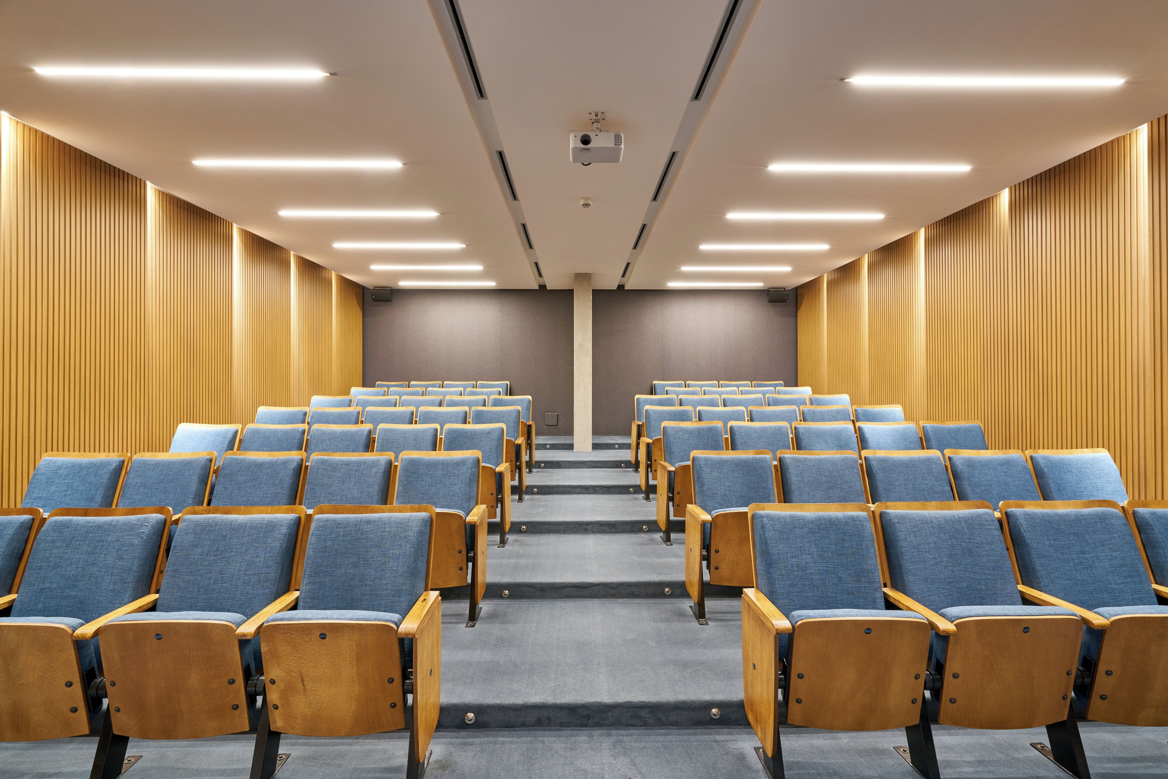 An auditorium with rows of blue chairs and wooden armrests, a grey carpet, and wooden panel walls.