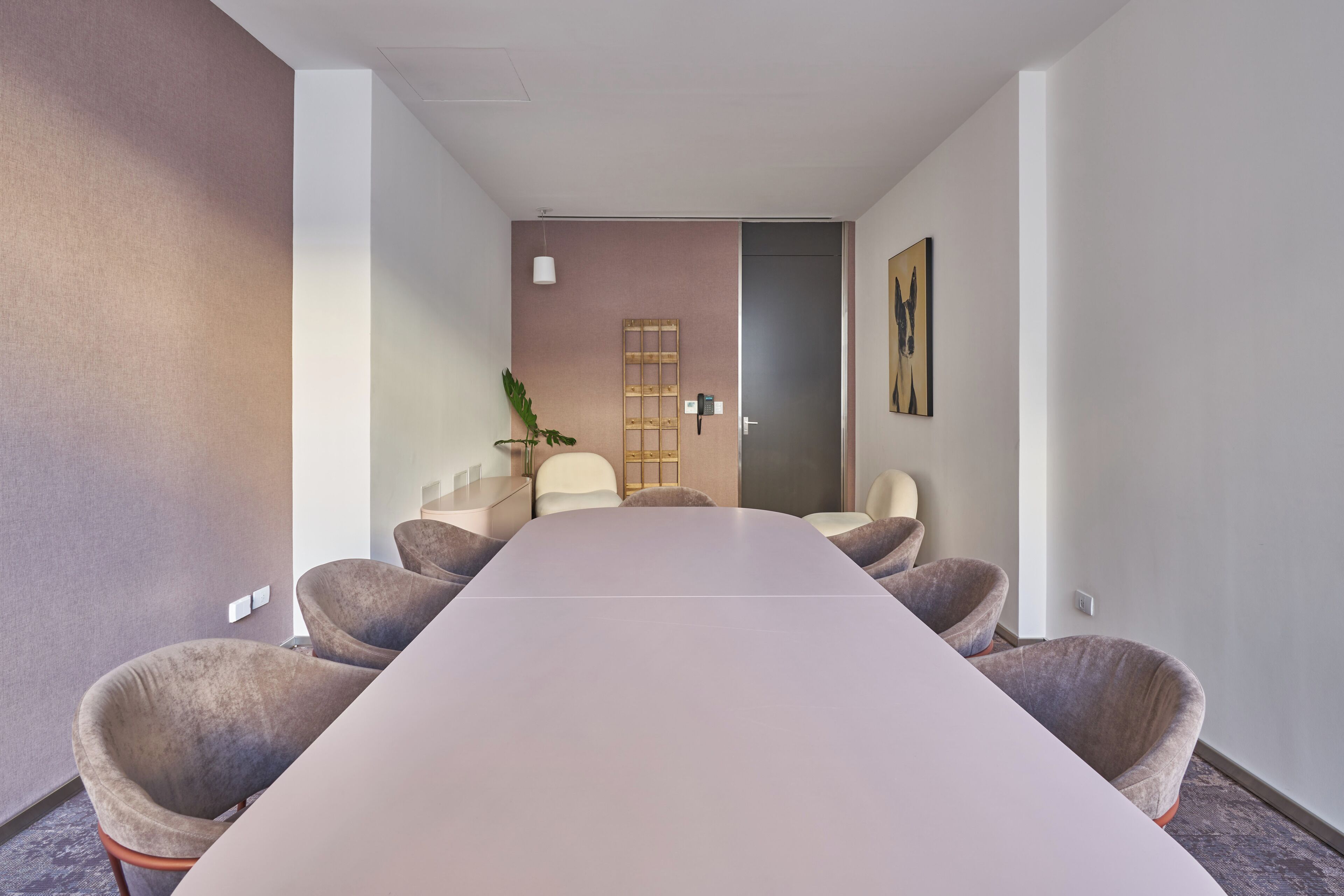 A conference room with a long table and six chairs, a plant, and a door in the background.