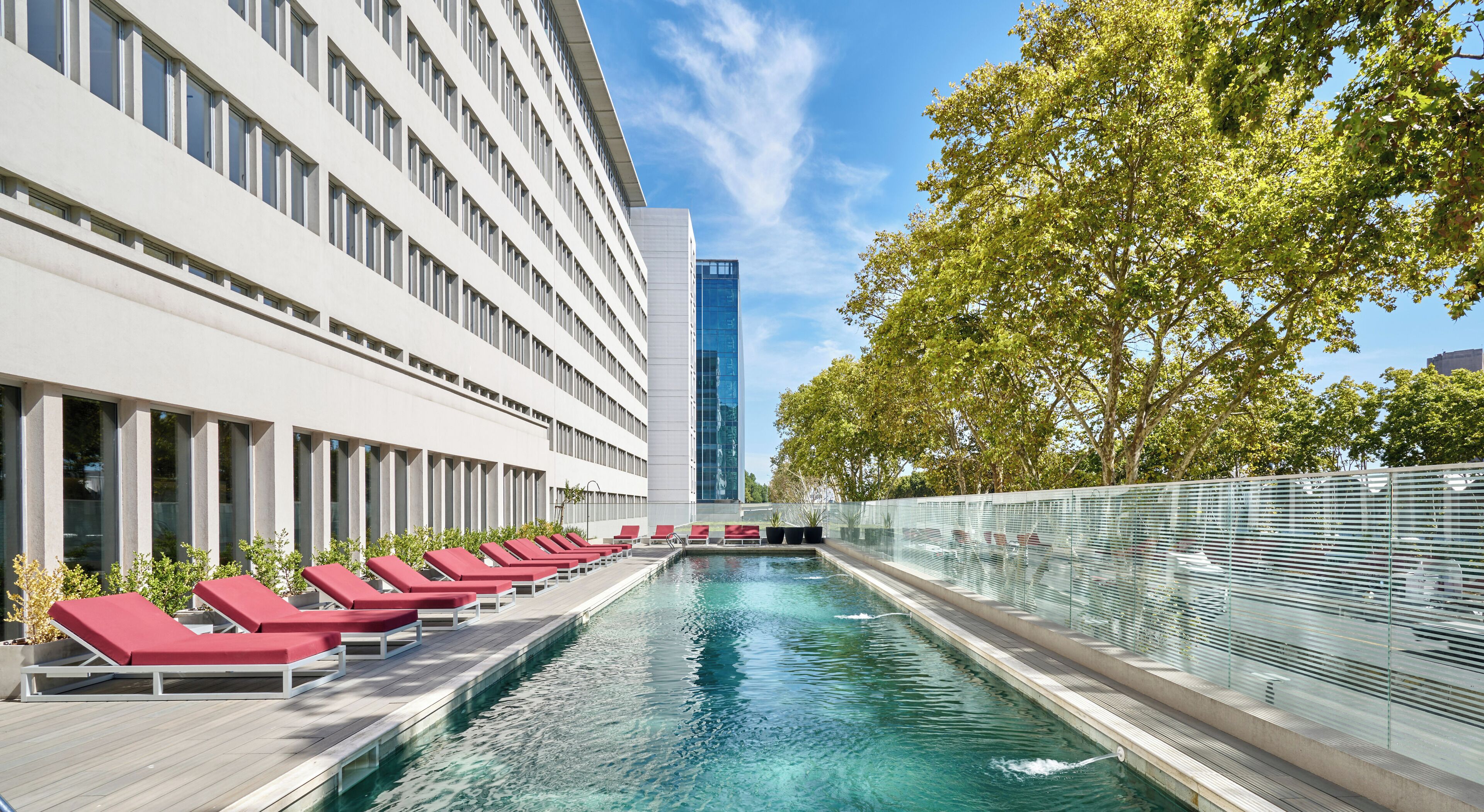 Un hotel moderno con piscina, sillones rojos y un edificio alto al fondo.