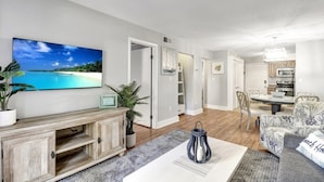 Ein Wohnzimmer mit großem Fernseher, einem Essbereich mit Tisch und Stühlen und einer Küche im Hintergrund.
