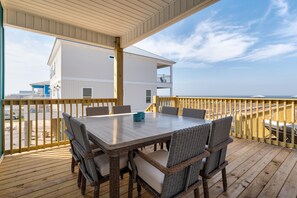 A covered outdoor deck with a dining table and wicker chairs, overlooking a beach and a white building. - Until Nextime - 2712 Bienville 5 Bedroom Home by RedAwning (Dauphin Island)