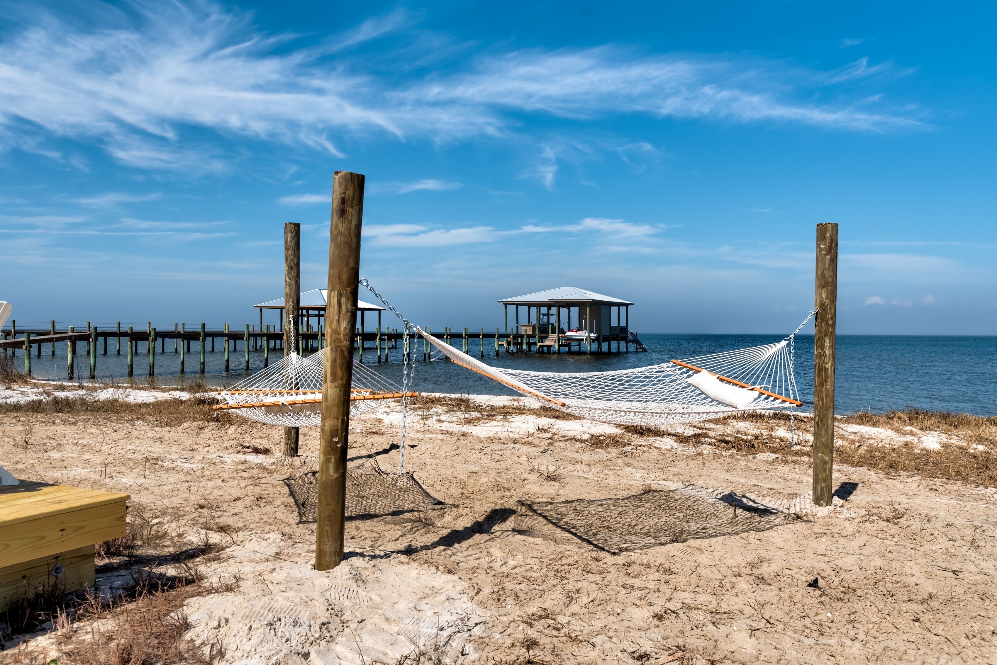 A beach with a hammock, wooden posts, a wooden structure, and a pier extending into the water.