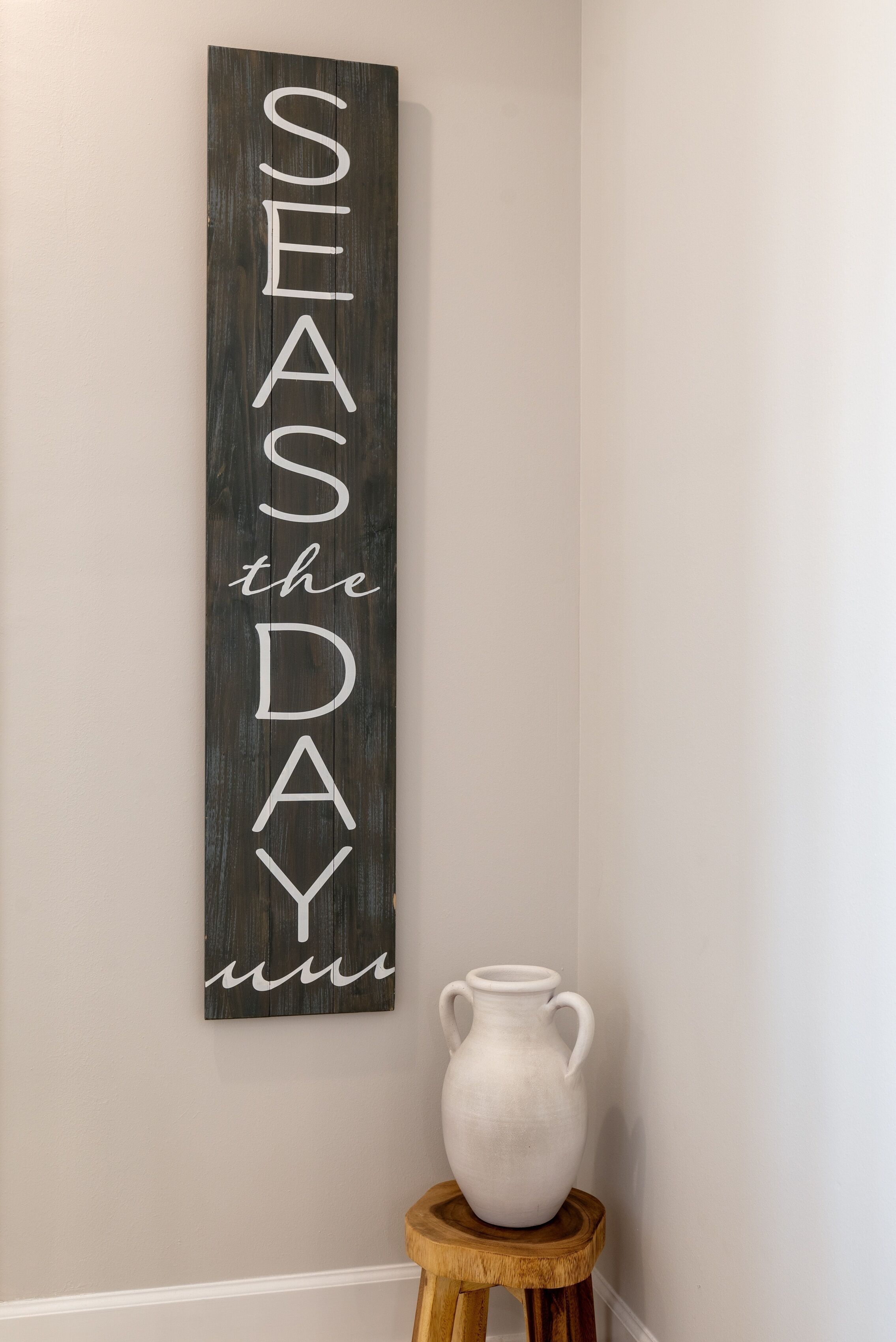 Um sinal de madeira vertical com as palavras 'SEASDAY' e 'the' numa parede, ao lado de um vaso branco sobre um banquinho de madeira.