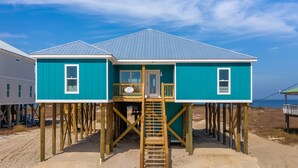 Una casa de playa de dos pisos con una terraza de madera y una escalera que conduce a la entrada.