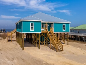 A two-story wooden structure with a staircase leading to the upper floor, situated on a sandy beach with a clear sky. - Until Nextime - 2712 Bienville 5 Bedroom Home by RedAwning (Dauphin Island)