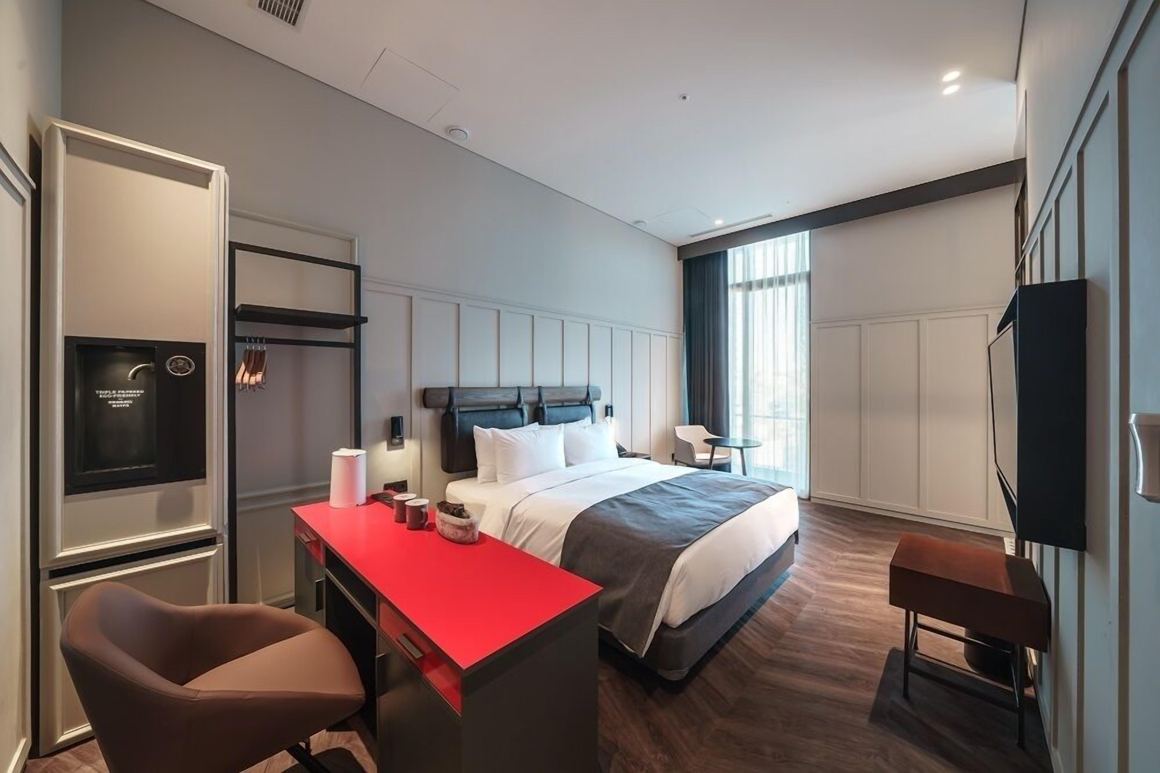Una habitación de hotel moderna con una cama, un escritorio con cubierta roja, una silla, un televisor y una ventana con cortinas.