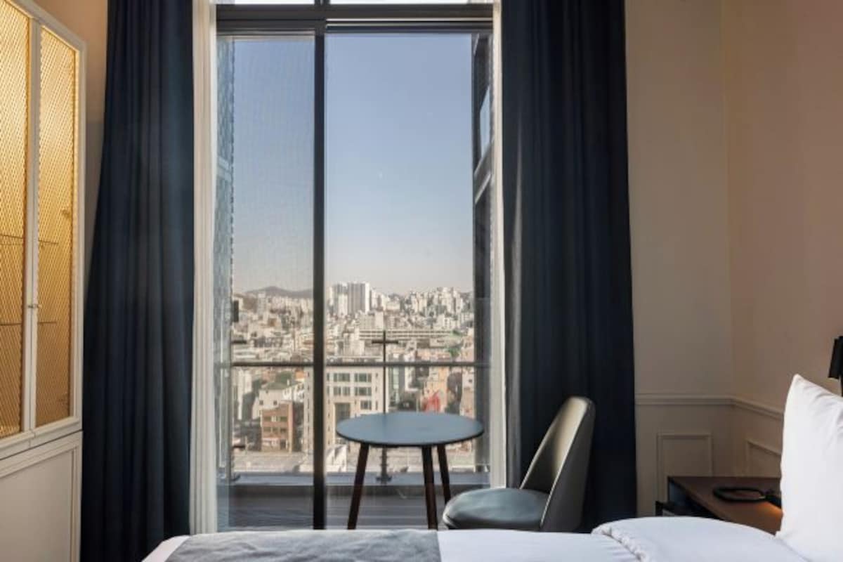 A hotel room with a large window, a bed, a chair, and a small table.