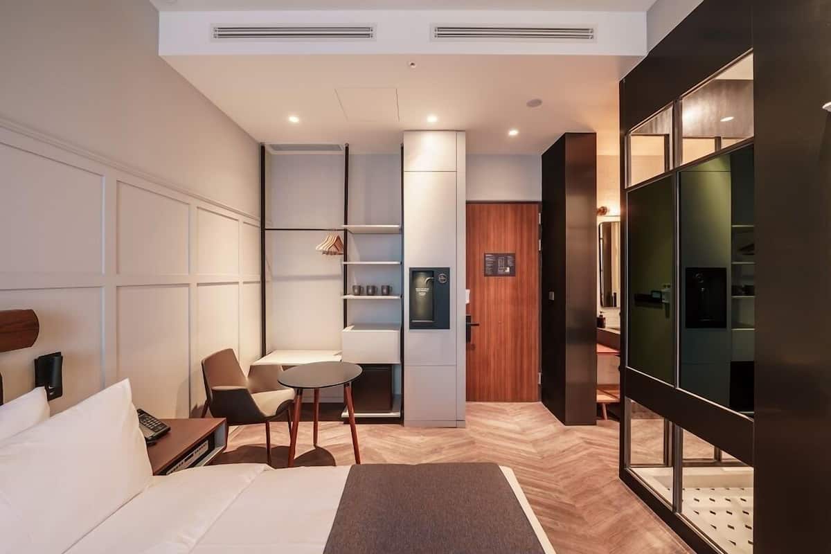 A modern hotel room with a bed, a desk, a chair, a small table, a wardrobe, and a glass partition.