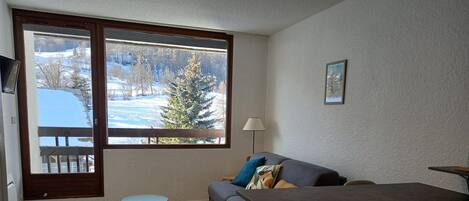 A cozy living room with a sofa, a small table, and a dining table. A large window offers a view of a snowy landscape.