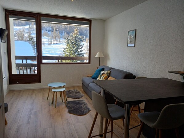 A cozy living room with a sofa, a small table, and a dining table. A large window offers a view of a snowy landscape.