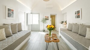 A modern living room with a sofa, a coffee table, and a hanging light fixture. - Amoramelia Suites (Santorini)