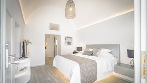 A modern bedroom with a large bed, bedside tables, a hanging light fixture, and a door leading to another room. - Amoramelia Suites (Santorini)