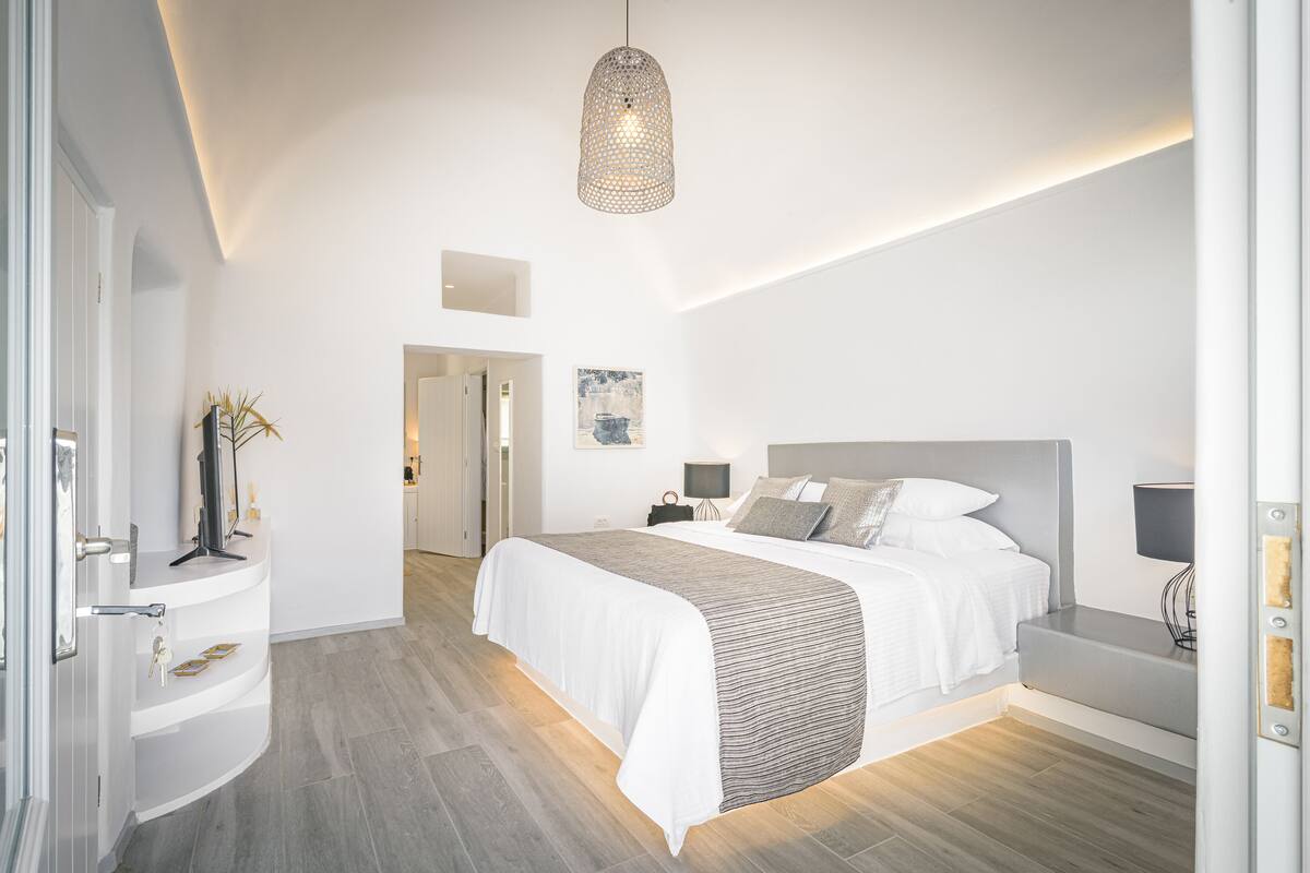 A modern bedroom with a large bed, bedside tables, a hanging light fixture, and a door leading to another room.