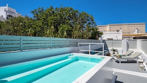 A rooftop pool with clear blue water, surrounded by white walls and a white fence. There are lounge chairs and a small table with a decorative item on the poolside. - Amoramelia Suites (Santorini)