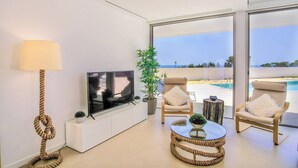 A modern living room with a flat-screen TV, a lamp, and a view of a pool and beach. - Ocean view Architectural Modern Villa (Luz)