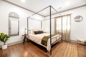 A modern bedroom with a four-poster bed, a bedside table, a potted plant, and a large mirror on the wall.