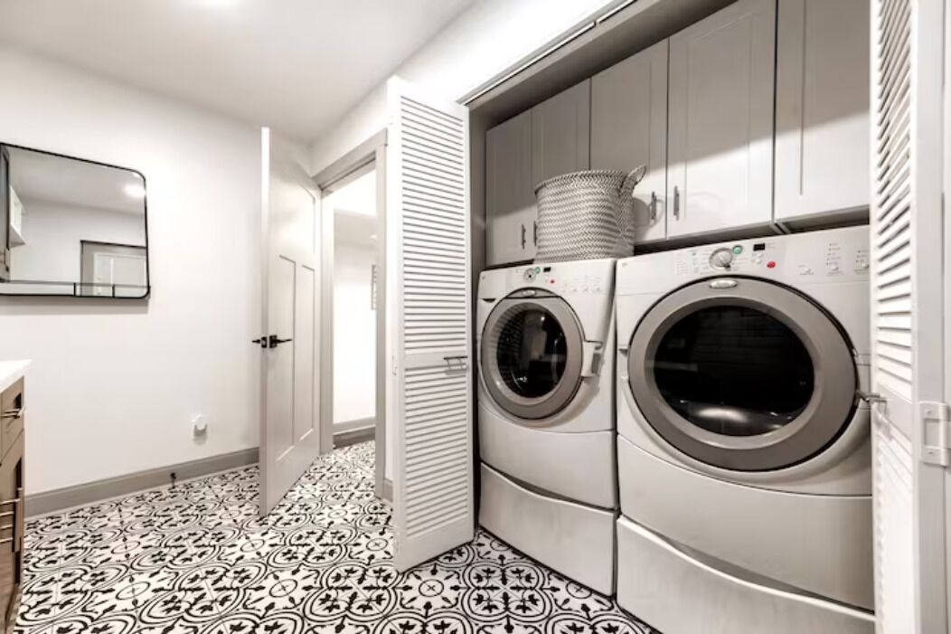 A laundry room with two stacked front-loading washing machines, a large mirror, and patterned floor tiles.